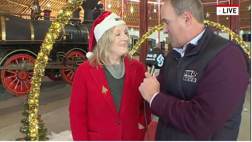 A reporter from WBFF interviews Executive Director Kris Hoellen. They are standing beneath golden arches covered with Christmas lights.